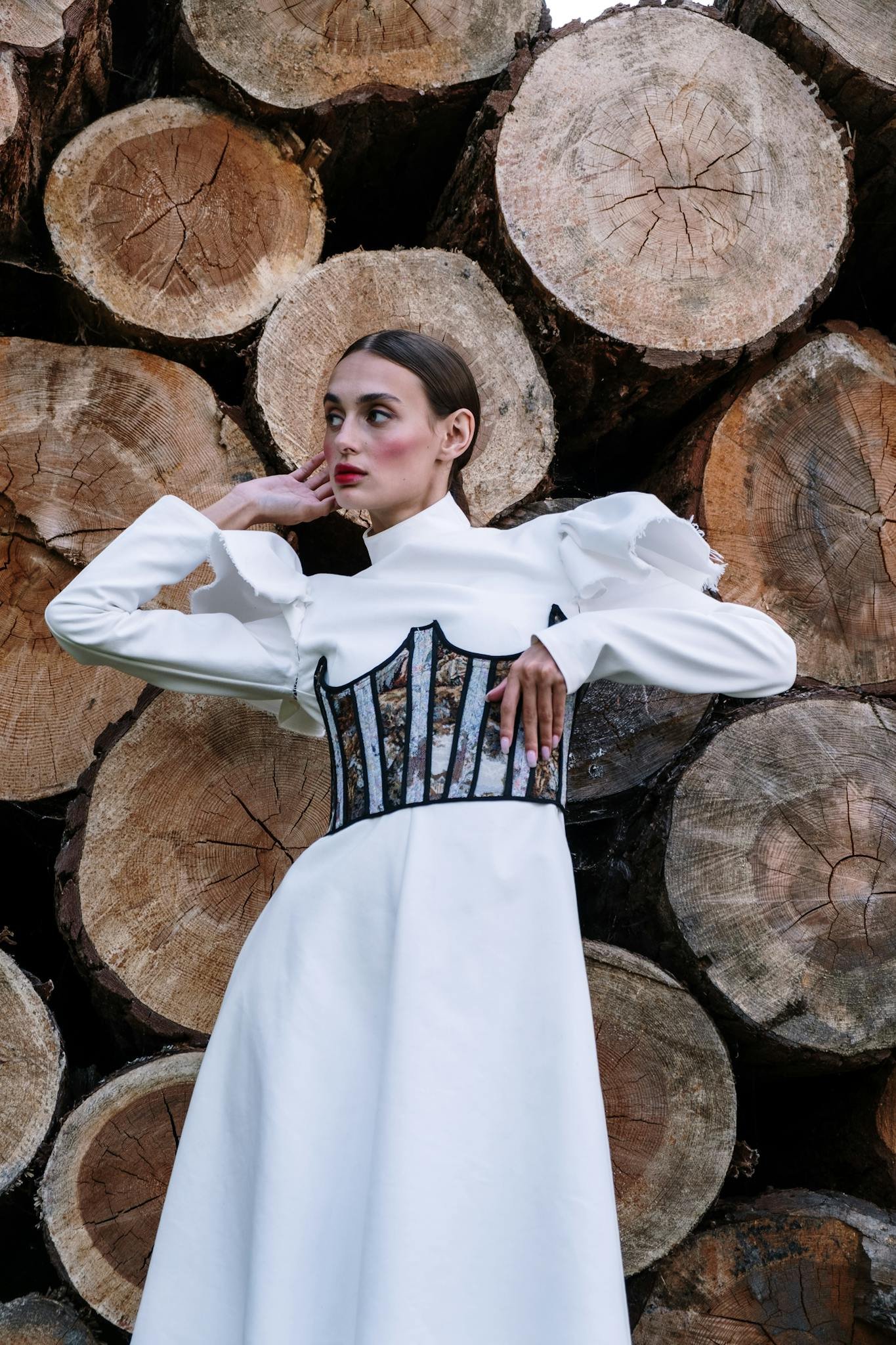 Stylish woman in a white dress posing confidently against a backdrop of wooden logs.